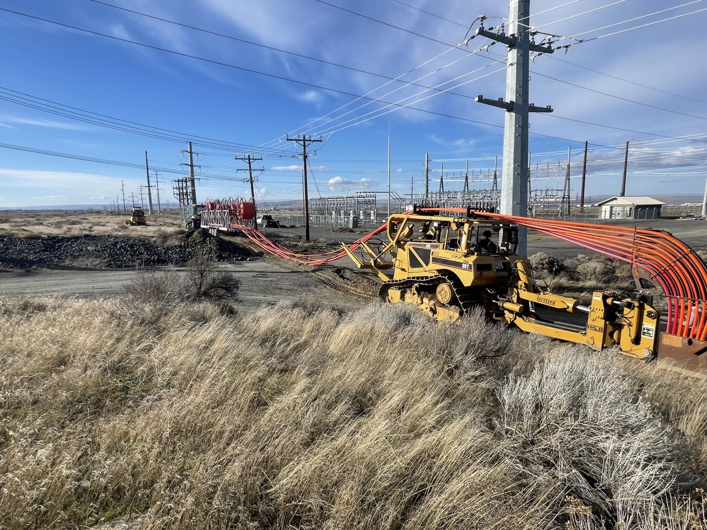 Cable plough drawing orange conduit across rangeland beside a substation