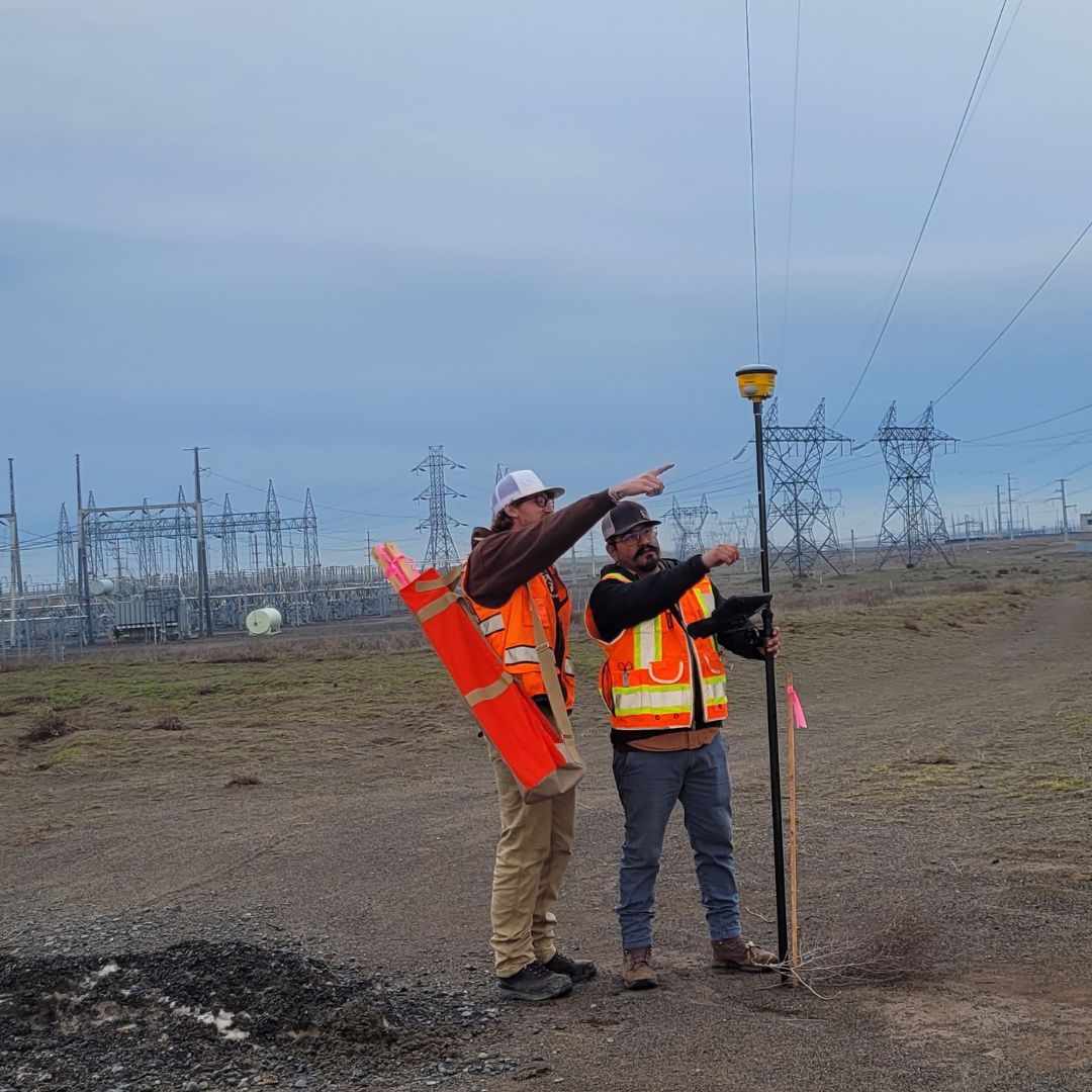 Two Windwave surveyors sighting a line with a survey rod beside a substation